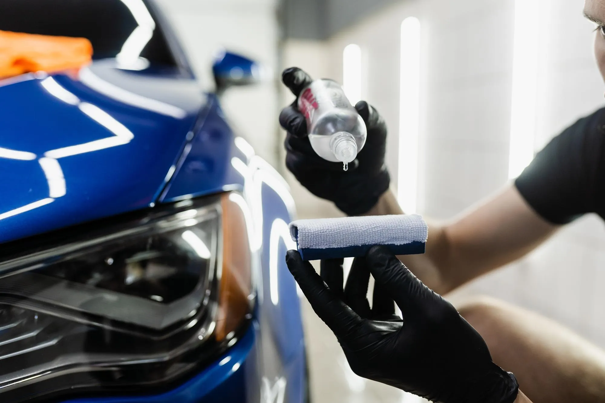 A technician from A1 Mobile Detailing & Ceramic, wearing black gloves, applies droplets of a liquid nano ceramic coating product onto a suede applicator pad, preparing to treat the glass surface of a glossy blue car.