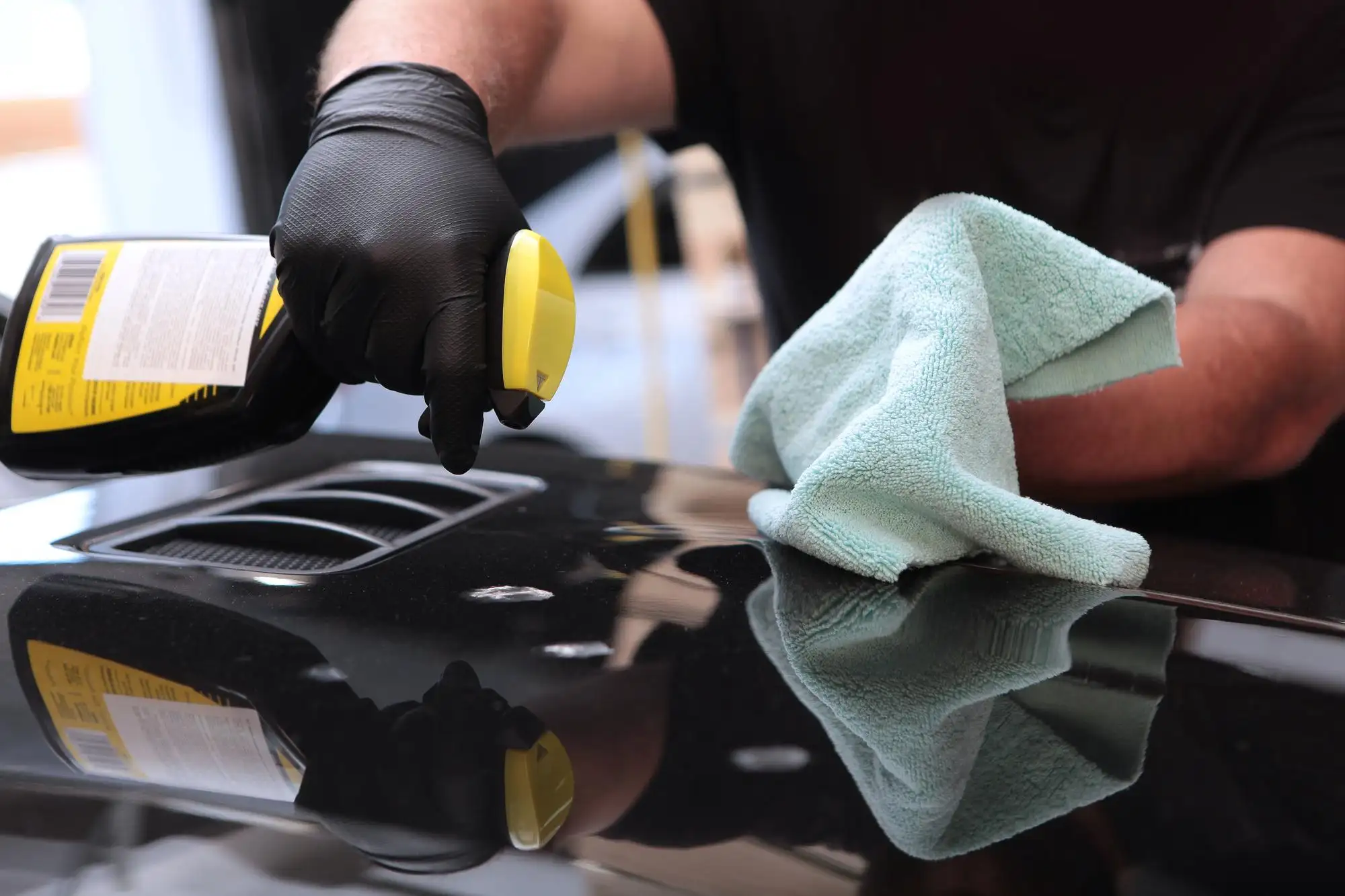 A professional at A1 Mobile Detailing & Ceramic performing Exterior Car Detailing by spraying a finishing agent onto a glossy black car hood and preparing to wipe with a microfiber towel.
