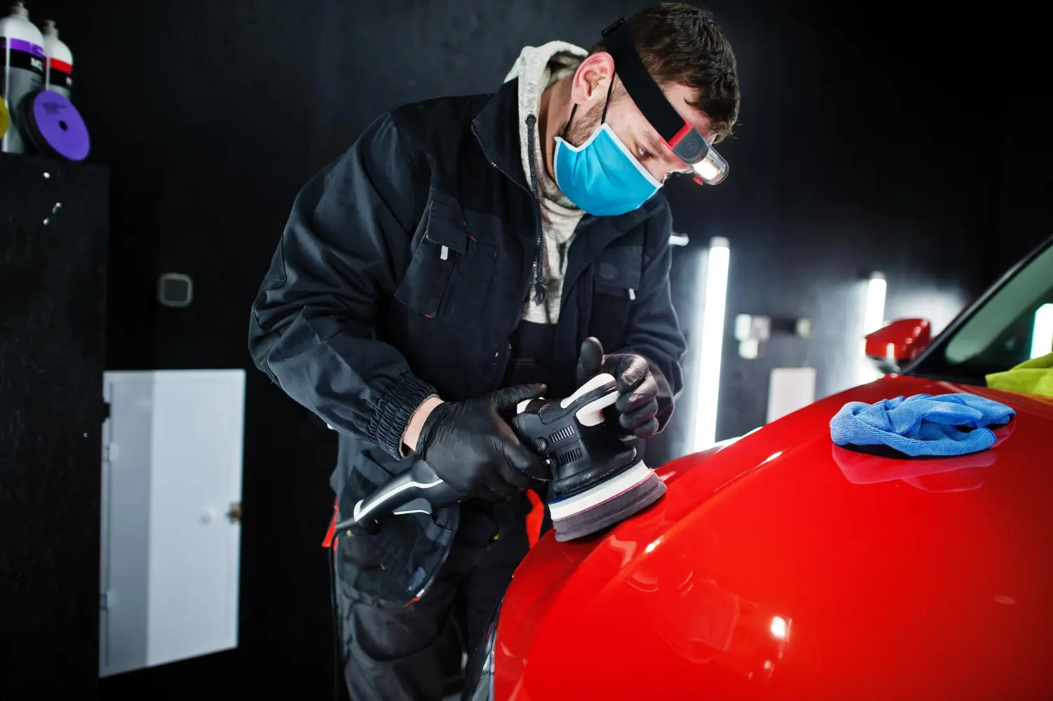 A detailer wearing a face mask and headlamp performing professional car paint correction on a red car with a dual-action polisher.